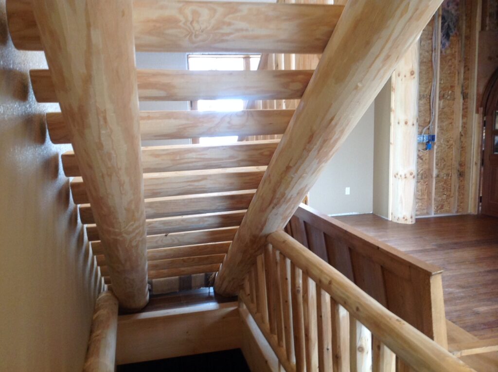 Access to a basement is visible from underneath the stairs, which is made entirely from untreated, light colored logs.